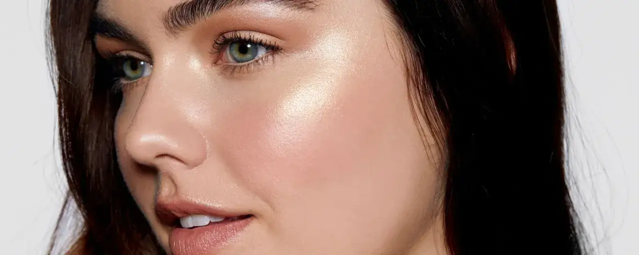 En kvinna med gr&ouml;na &ouml;gon och m&ouml;rkt h&aring;r som har f&aring;tt en gl&ouml;dande finish genom att applicera highlighter p&aring; kindbenen.