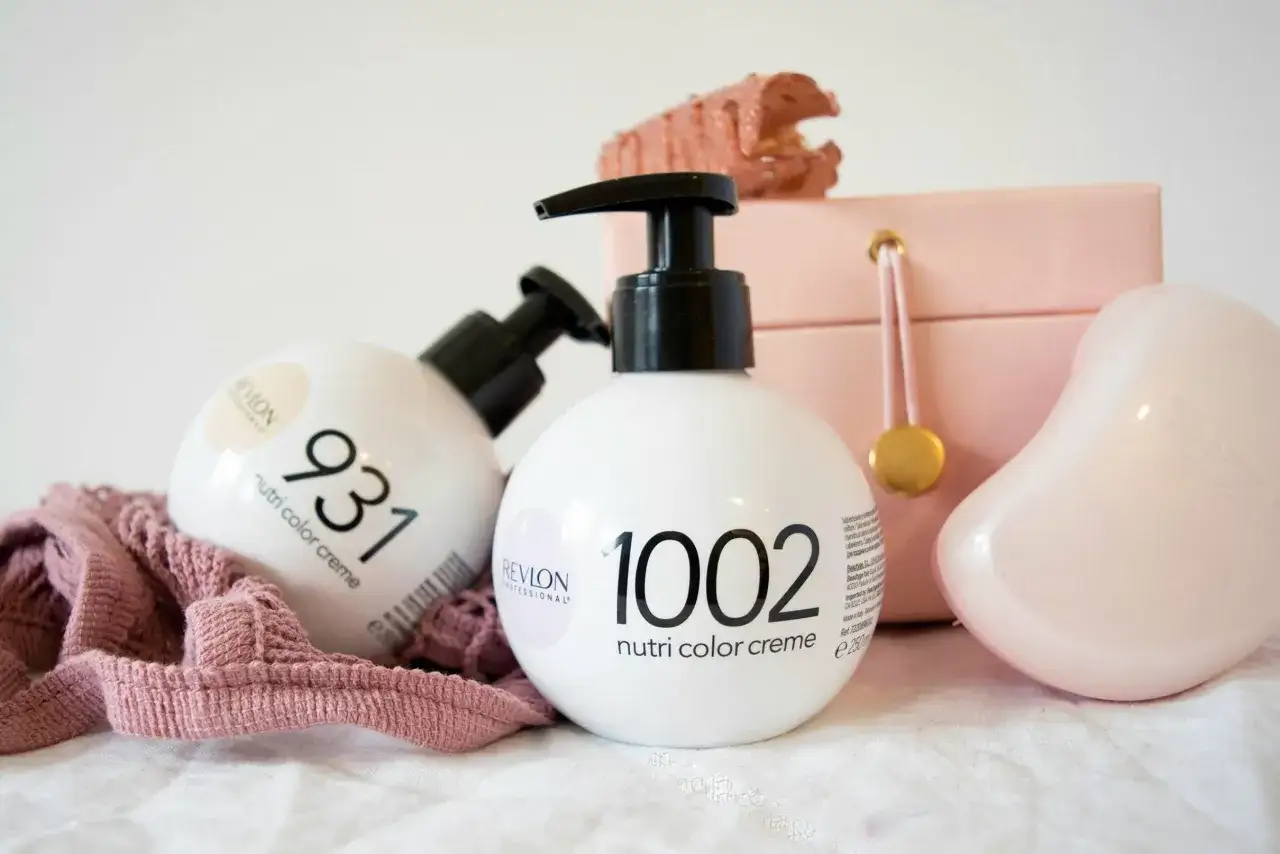 Tv&aring; flaskor Revlon h&aring;rf&auml;rg, Nutri Color Creme, st&aring;r bredvid en rosa necess&auml;r och en stickad tygbit.